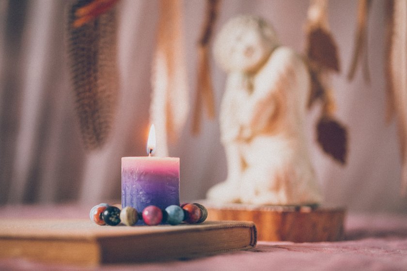 Buddha of compassion figurine with a book, a lighted candle standing in a pearl bracelet in front of the part of a defocused dreamcatcher in soft rose ambience for mindfulness and wisdom concepts.  Selective focus and added grain. Part of a series.